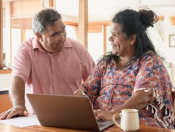 2 people having a conversation in their home. One of them is typing on a laptop.