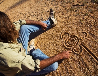 An Aboriginal person drawing art in the dirt on the ground.